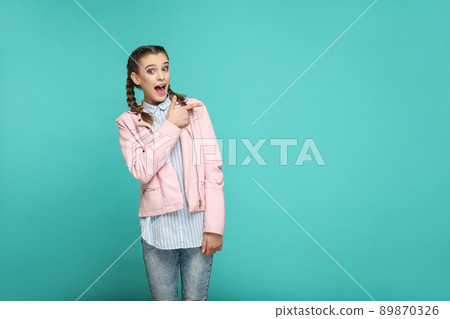 surprised face pointing copyspace portrait of beautiful cute girl standing with makeup and pigtail hairstyle in striped blue shirt pink jacket. indoor, studio shot isolated on blue or green background surprised face pointing copyspace portrait of beautiful cute girl standing with makeup and pigtail hairstyle in striped blue shirt pink jacket. indoor, studio shot isolated on blue or green background 89870326