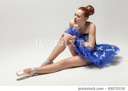 knee pain. ballerina with bun collected hair wearing blue dress and pointe shoes holding on injured knee while sitting on white floor. indoor, studio shot. 89870330