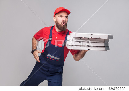 Hurry up! Young amazed delivery man with beard in blue uniform and red t-shirt holding stack pizza boxes and running to delivery on grey background. Indoor, studio shot, isolated, copy space. 89870331
