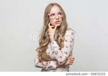 Thinking concept. Puzzlement long haired blonde woman in pink eyeglasses, touching her chin and looking up. Studio shot. Isolated on gray background 89870332