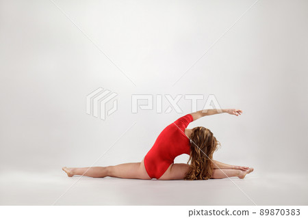 young athletic blonde woman in red leotard sitting on string. hanumanasana yoga pose. isolated indoor studio shot on light gray background. healthy lifestyle and leisure activity concept. 89870383