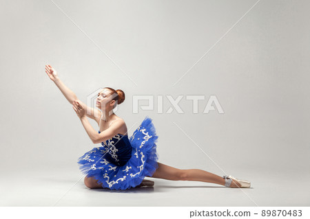 attractive ballerina with bun collected hair wearing blue dress and pointe shoes graceful sitting on white studio floor. indoor, studio shot. attractive ballerina with bun collected hair wearing blue dress and pointe shoes graceful sitting on white studio floor. indoor, studio shot. 89870483