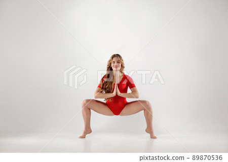 attractive athletic curly blonde woman with makeup in red leotard doing yoga pose .isolated indoor studio shot on light gray background. healthy lifestyle and leisure activity concept. 89870536