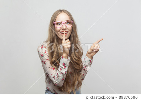 Happiness beautiful young girl, showing finger at copy space and looking at camera with conspiracy sign. Studio shot, isolated on gray background Happiness beautiful young girl, showing finger at copy space and looking at camera with conspiracy sign. Studio shot, isolated on gray background 89870596