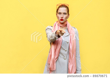 The beautiful woman, wearing autumn coat, opening mouths widely, having surprised shocked looks, pointing finger at camera. Studio shot, yellow wall The beautiful woman, wearing autumn coat, opening mouths widely, having surprised shocked looks, pointing finger at camera. Studio shot, yellow wall 89870671