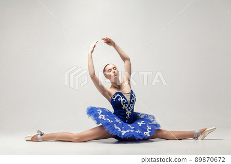 attractive ballerina with bun collected hair wearing blue dress and pointe shoes sitting on string isolated on white background. indoor, studio shot. 89870672