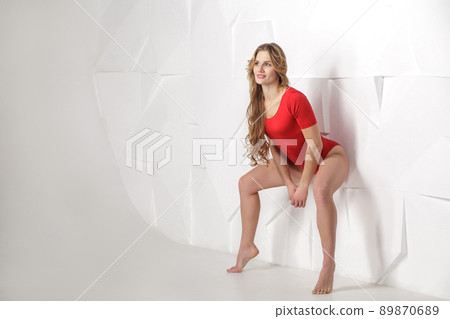 beautiful sexy girl in red bodysuit with long curly blonde hairstyle and makeup and perfect body lean and posing on white wall and looking away. good emotions and feeling concept. indoor studio shot. 89870689