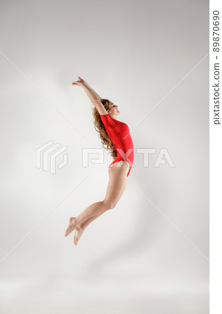 Happy beautiful blonde gymnast with curly long hair in red leotard jumping. indoor studio shot on light gray background. healthy lifestyle and leisure activity concept. 89870690