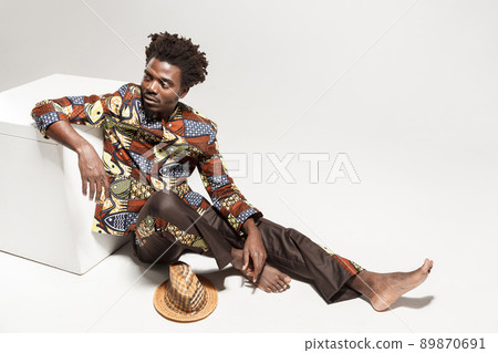 Wondering afro man in traditional national clothes sit on cube. Indoor, isolated on gray background Wondering afro man in traditional national clothes sit on cube. Indoor, isolated on gray background 89870691