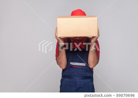 Portrait of young logistic delivery man with beard in blue uniform and red t-shirt standing and covering face with the cardboard box on grey background. Indoor, studio shot, isolated, copy space Portrait of young logistic delivery man with beard in blue uniform and red t-shirt standing and covering face with the cardboard box on grey background. Indoor, studio shot, isolated, copy space 89870696