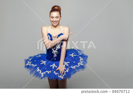 Portrait of happy beautiful ballerina woman in blue costume with makeup standing against gray background and pointing at gray wall. emotion and expression concept. indoor studio shot. Portrait of happy beautiful ballerina woman in blue costume with makeup standing against gray background and pointing at gray wall. emotion and expression concept. indoor studio shot. 89870712