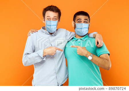 Portrait of two young worker men with surgical medical mask standing, hugging, showing and pointing each other and looking at camera with funny face. indoor studio shot isolated on orange background. Portrait of two young worker men with surgical medical mask standing, hugging, showing and pointing each other and looking at camera with funny face. indoor studio shot isolated on orange background. 89870724