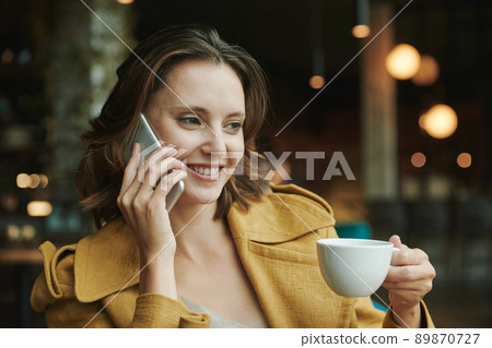 Confident young man in casual shirt sitting in armchair and looking away while talking by phone in modern cafe Confident young man in casual shirt sitting in armchair and looking away while talking by phone in modern cafe 89870727