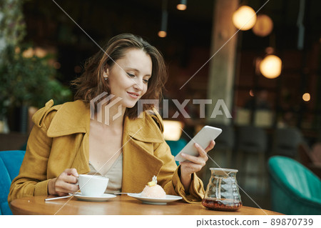 Young bearded manager sitting at table with coffee cup and croissant and recording voice message for colleague using smartphone in cafe 89870739