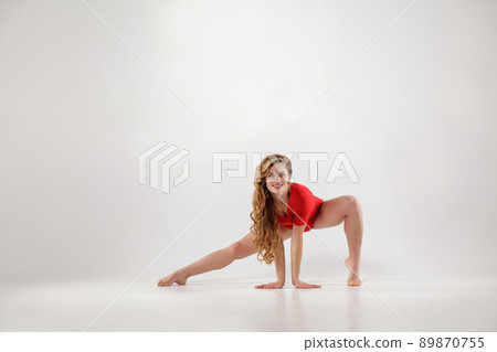 attractive athletic blonde woman with curly long hair and makeup in red leotard doing yoga pose. isolated indoor studio shot on light gray background. healthy lifestyle and leisure activity concept. 89870755