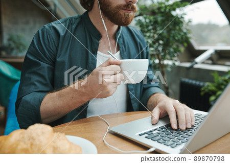 Young bearded manager sitting at table with coffee cup and croissant and recording voice message for colleague using smartphone in cafe Young bearded manager sitting at table with coffee cup and croissant and recording voice message for colleague using smartphone in cafe 89870798
