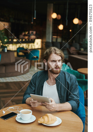 Serious handsome guy with beard sitting at table in cafe and drinking coffee while reading mail on smartphone Serious handsome guy with beard sitting at table in cafe and drinking coffee while reading mail on smartphone 89870823