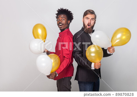 Two shocked friend wearing red shirt and black jacket holding many air balloon and looking at camera with shocked face. Indoor shot Two shocked friend wearing red shirt and black jacket holding many air balloon and looking at camera with shocked face. Indoor shot 89870828