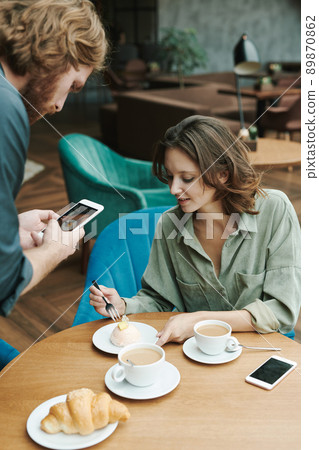 Busy young bearded man sitting at table in modern cafe and drinking coffee while discussing project by phone Busy young bearded man sitting at table in modern cafe and drinking coffee while discussing project by phone 89870862