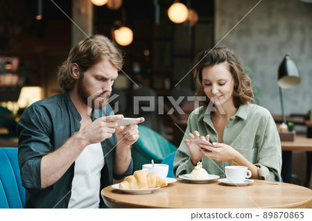 Concentrated young bearded man in casual shirt drinking coffee and eating croissant while checking smartphone in cafe Concentrated young bearded man in casual shirt drinking coffee and eating croissant while checking smartphone in cafe 89870865