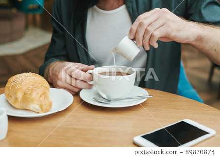 Busy young bearded man sitting at table in modern cafe and drinking coffee while discussing project by phone Busy young bearded man sitting at table in modern cafe and drinking coffee while discussing project by phone 89870873