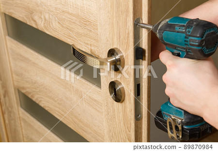 handyman repair the door lock in the room. man repairing the door handle furniture. carpenter at lock installation with electric drill into interior wood door handyman repair the door lock in the room. man repairing the door handle furniture. carpenter at lock installation with electric drill into interior wood door 89870984