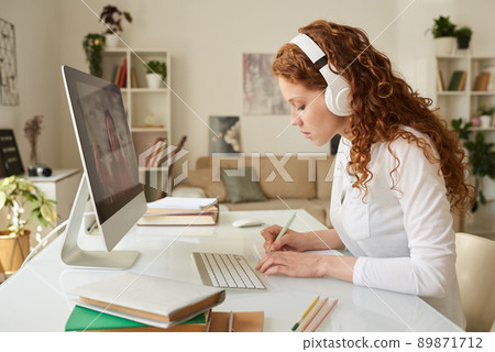 Concentrated curly-haired student girl in headphones sitting at table and writing down information said by online tutor Concentrated curly-haired student girl in headphones sitting at table and writing down information said by online tutor 89871712