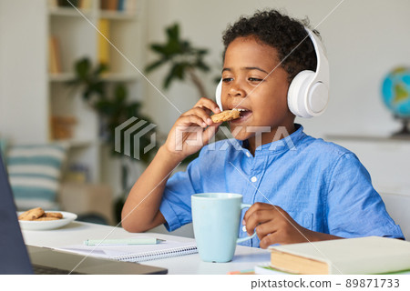 Cute African-American boy in wireless headphones sitting at desk and eating cookie while watching educational lecture Cute African-American boy in wireless headphones sitting at desk and eating cookie while watching educational lecture 89871733
