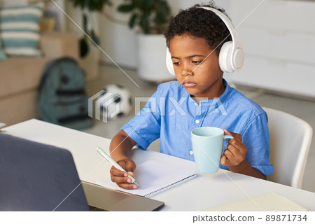 Thoughtful African-American schoolboy in blue shirt sitting at desk and drinking tea while listening to online lesson Thoughtful African-American schoolboy in blue shirt sitting at desk and drinking tea while listening to online lesson 89871734