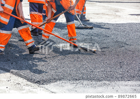 A team of road workers in orange overalls level hot asphalt with metal levels and shovels on a sunny day. 89872665