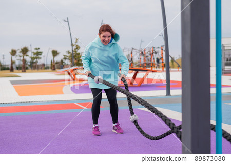 Caucasian woman in a mint sweatshirt is training with battle ropes at the sports ground.  89873080