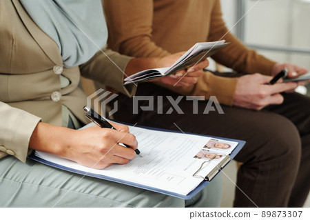 Unrecognizable Muslim woman in hijab sitting on chair in visa service agency filling in application form while waiting for appointment 89873307