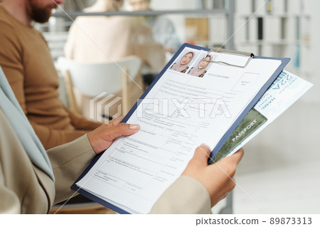 Unrecognizable Muslim woman wearing hijab sitting on chair waiting for appointment in queue holding visa application form and other documents 89873313