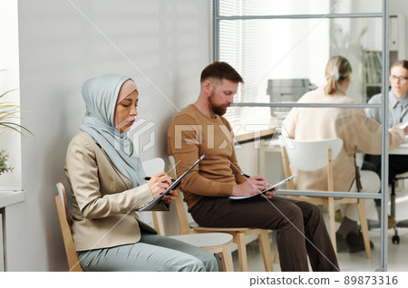 Caucasian man and Muslim woman in hijab sitting on chairs in embassy or visa service agency filling in application forms while waiting in queue 89873316