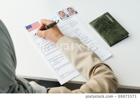 High angle view of unrecognizable Muslim woman sitting at desk with passport on it filling in visa application form 89873371
