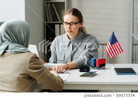 Modern Caucasian consular officer sitting at desk in front of Muslim woman explaining her how to fill in application form 89873374
