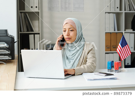 Portrait of young adult Muslim woman wearing hijab working as consular officer in US embassy texting on laptop and talking on phone 89873379