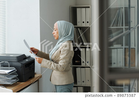 Side view of modern Muslim woman wearing hijab working in office standing in front of printer checking copies of document Side view of modern Muslim woman wearing hijab working in office standing in front of printer checking copies of document 89873387