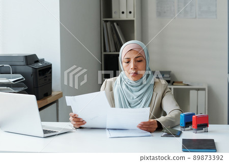 Modern Muslim woman wearing hijab sitting at desk in office doing paperwork checking information correctness 89873392