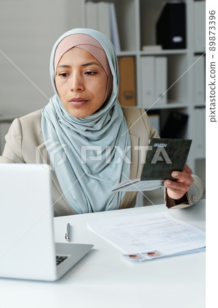 Vertical portrait of young adult Muslim woman wearing hijab sitting at desk in consulate office working with documents using laptop 89873396