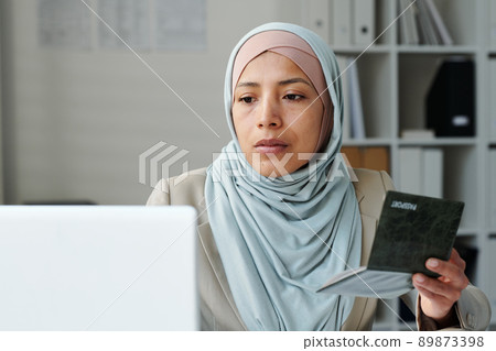 Portrait of serious young adult Muslim woman wearing hijab sitting in embassy office using laptop to work with documents 89873398