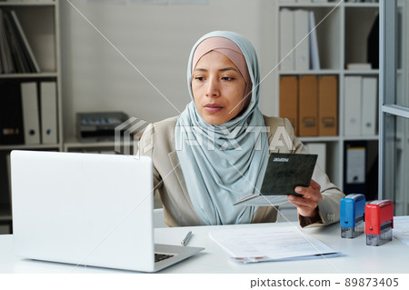 Young adult Muslim woman wearing hijab sitting at desk in embassy office working with visa applicants documents using laptop Young adult Muslim woman wearing hijab sitting at desk in embassy office working with visa applicants documents using laptop 89873405