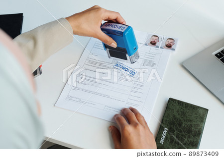 High angle view of unrecognizable Muslim woman wearing hijab working as consular officer sitting at desk putting approved stamp on document paper High angle view of unrecognizable Muslim woman wearing hijab working as consular officer sitting at desk putting approved stamp on document paper 89873409