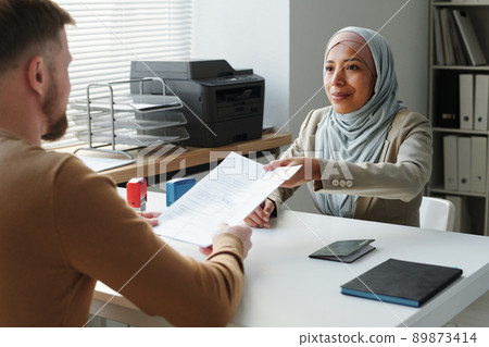 Modern Muslim woman wearing hijab working in embassy sitting at desk in front of Caucasian man giving him documents after visa approval 89873414