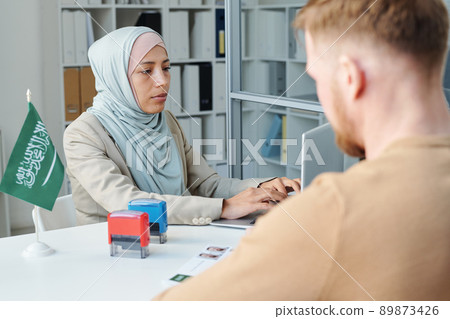 Modern Muslim woman working in Saudi Arabia embassy consulting unrecognizable man for visa application 89873426