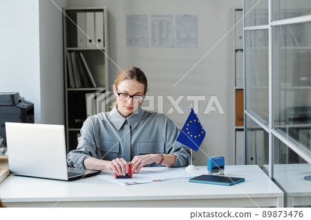Caucasian woman working as consular officer in embassy sitting at desk in office putting rejection and approval seals on visa applications 89873476