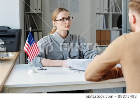 Caucasian woman wearing eyeglasses working in US embassy interviewing unrecognizable man for visa 89873498