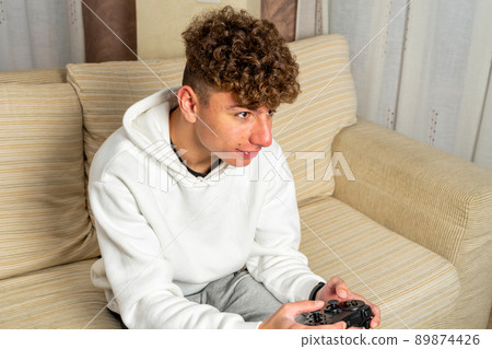 Portrait of attractive dark-haired Caucasian man, holding joystick and playing video games on TV on vacation, sits at home on cozy sofa, is successful, is winning in crazy car race. 89874426