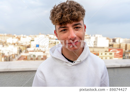 Portrait of smiling young Caucasian man with curly hair dressed in a white sweatshirt looking at the camera with the city skyline in the background. Portrait of smiling young Caucasian man with curly hair dressed in a white sweatshirt looking at the camera with the city skyline in the background. 89874427
