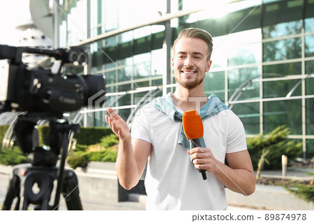 Young male journalist with microphone working on city street Young male journalist with microphone working on city street 89874978